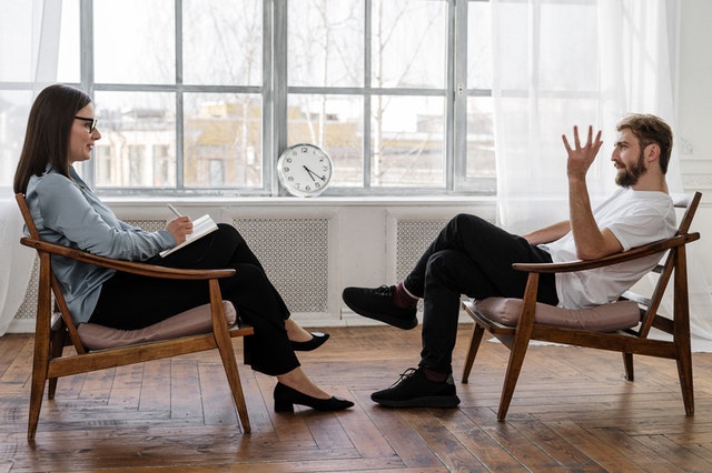 A man sitting in a reclining wooden chair, facing a woman in a similar chair, at a therapy session. The man is making an animated hand gesture, and the woman is taking notes.