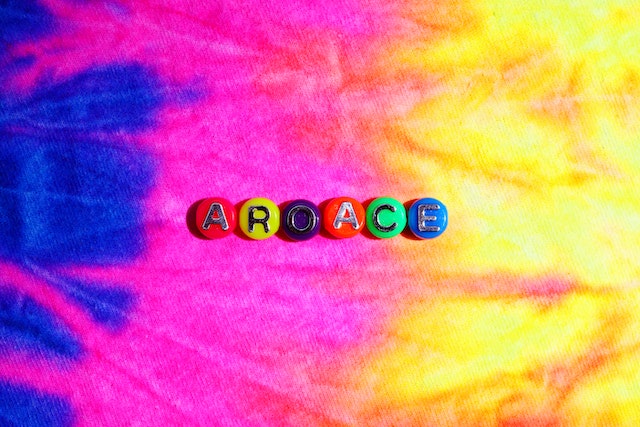 Purple, pink, and yellow tie-dye background with the word aroace laid out across the center in round colored pins.