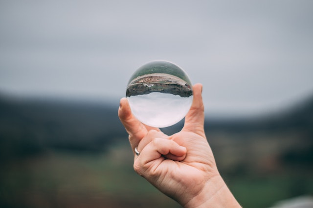 Very blurry photo of a landscape and the sky, with a white person's hand holding up a lens that brings the landscape into focus in mirror image.