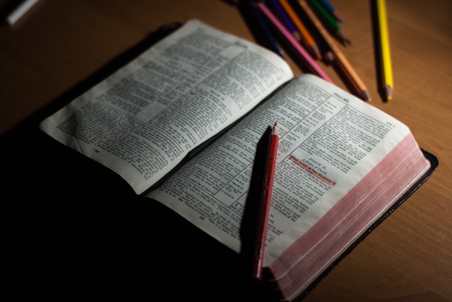 Open Bible resting on a wood surface with a highlighter sitting on it next to a highlighted line of text, and more writing instruments scattered on the table behind it.