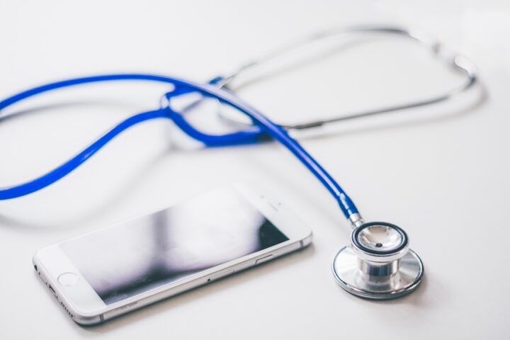 A white metallic background with a cell phone sitting in the lower left corner, and a stethoscope with blue tubing occupying the rest of the space