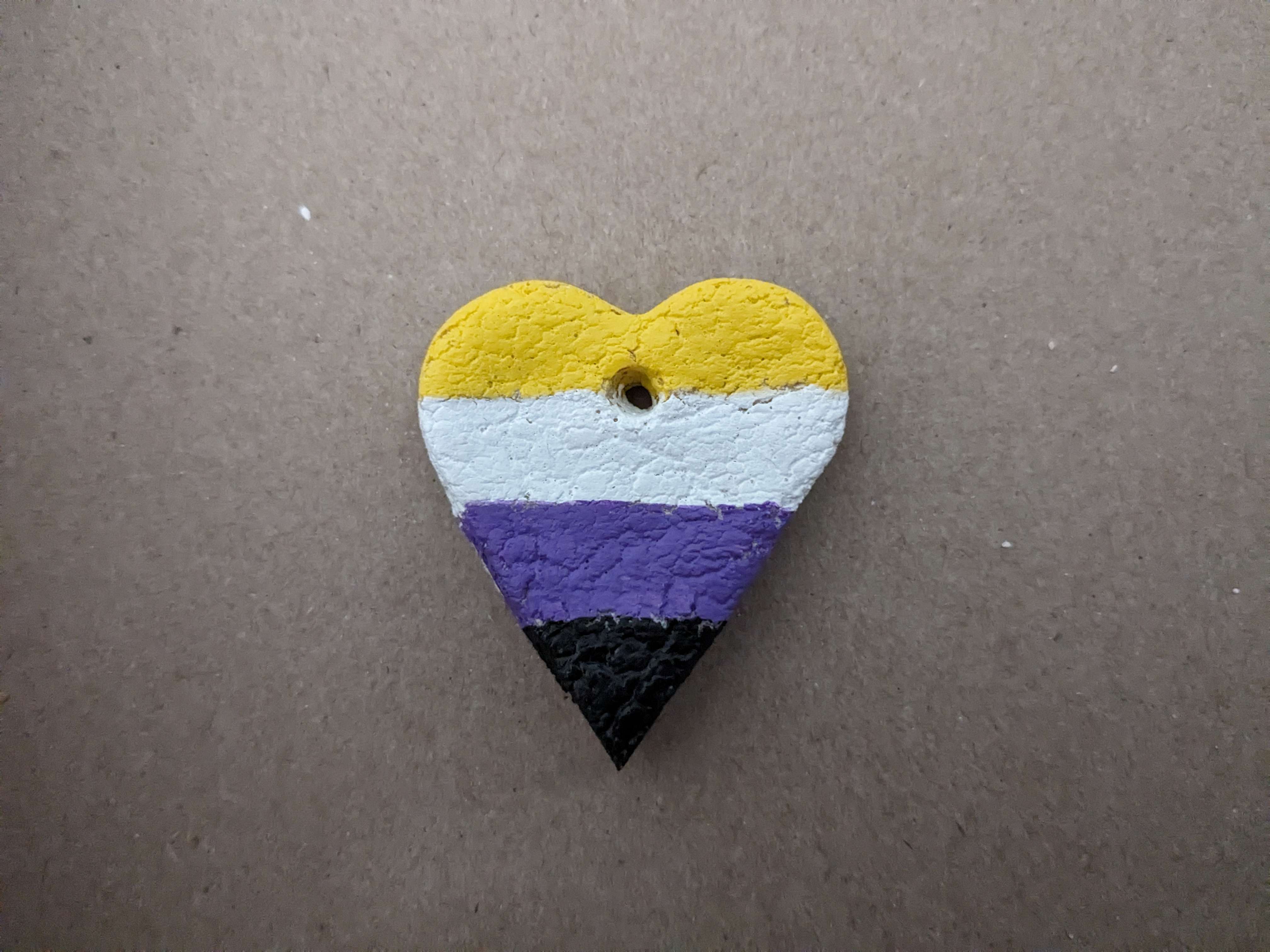Heart ornament painted like the nonbinary flag sitting on a thin cardboard backdrop.