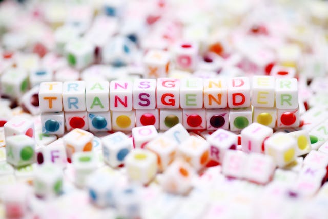 Square letter beads spelling out the word transgender, sitting among a pile of other square letter beads scattered on a white surface.